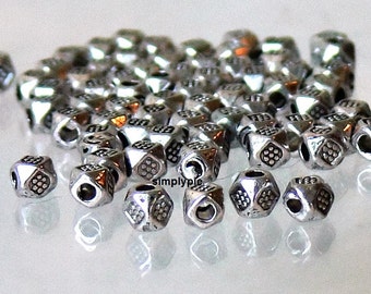 May include: A collection of small, silver-colored metal beads. Each bead is cube-shaped with a geometric pattern on each face. The beads are scattered across a reflective surface, creating a shimmering effect. The beads are likely for jewelry making.