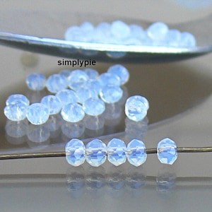 May include: Close-up of iridescent, faceted, light blue beads. Some beads are loose, while others are strung on a thin wire. The beads have a translucent quality, reflecting light. The text "simplypie" is visible.
