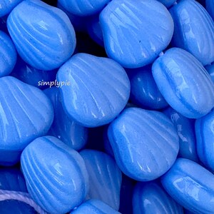 May include: A close-up shot of a pile of light blue, shell-shaped beads. Each bead has a glossy finish and is adorned with curved, ridged detailing. The beads are scattered, creating a textured and visually appealing arrangement.