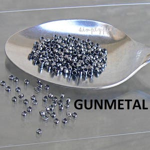 Puede incluir: Primer plano de una cuchara de plata llena de peque&ntilde;as cuentas redondas de color gris oscuro. Las cuentas est&aacute;n esparcidas sobre una superficie gris claro con la palabra "GUNMETAL" impresa en negro.