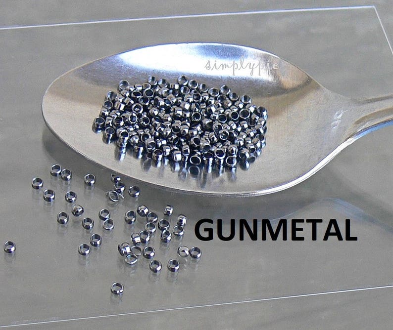 Puede incluir: Primer plano de una cuchara de plata llena de peque&ntilde;as cuentas redondas de color gris oscuro. Las cuentas est&aacute;n esparcidas sobre una superficie gris claro con la palabra "GUNMETAL" impresa en negro.