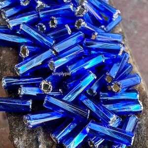 May include: A collection of vibrant blue glass beads. Each bead is a small, rectangular tube with a metallic silver-colored cap on each end. The beads are scattered across a textured surface, creating a sparkling effect. The word "simplypie" is visible.