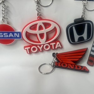 May include: A set of six keychains featuring different car logos. The keychains are made of red, white, black, and blue plastic. The logos include Nissan, Toyota, Honda, and Mitsubishi.