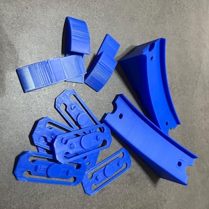 Puede incluir: Una colección de componentes de plástico azul brillante impresos en 3D. El surtido incluye piezas curvas con agujeros, piezas rectangulares con crestas y piezas más pequeñas y planas con detalles circulares y ovalados. Los objetos están dispersos sobre una superficie gris.