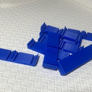 Könnte beinhalten: Eine Ansammlung hellblauer, rechteckiger Kunststoffkomponenten. Einige sind in Reihen angeordnet, andere sind verstreut. Die Teile haben ein einfaches, geometrisches Design und liegen auf einem weißen, strukturierten Hintergrund.