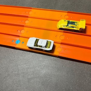 May include: Two toy race cars, one white and one yellow, are positioned on an orange race track. The white car has a yellow tinted windscreen. The yellow car has black accents and a rear spoiler. The track is on a grey surface.