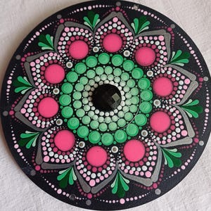 Pu&ograve; includere: Un mandala circolare nero con un motivo a punti verdi e rosa. Il centro del mandala ha un punto nero con una forma di diamante.