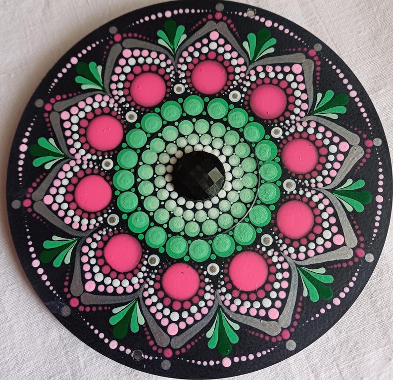 Pu&ograve; includere: Un mandala circolare nero con un motivo a punti verdi e rosa. Il centro del mandala ha un punto nero con una forma di diamante.