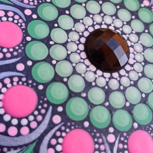 Pu&ograve; includere: Una pietra nera con un design a mandala verde, bianco e rosa. Il centro del mandala ha una pietra preziosa nera.