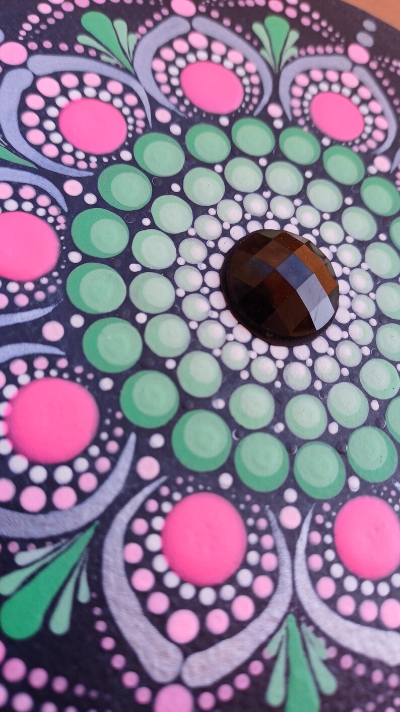 Pu&ograve; includere: Una pietra nera con un design a mandala verde, bianco e rosa. Il centro del mandala ha una pietra preziosa nera.
