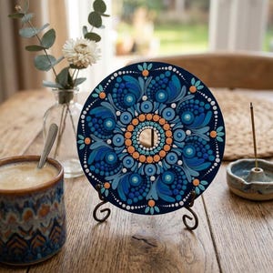 Peut inclure: Un disque circulaire orné d'un motif mandala peint à la main dans des tons bleu foncé, bleu clair et pêche. L'œuvre se compose de motifs de points complexes et de formes de feuilles tourbillonnantes disposées symétriquement autour d'une découpe centrale.