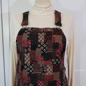 May include: A patchwork overall dress in shades of brown, red, and black. The dress features a variety of geometric patterns and textures. It has adjustable straps with metal hardware and is worn over a cream-colored turtleneck.