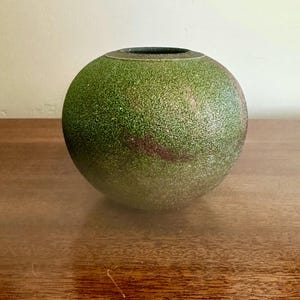 May include: A spherical ceramic vase with a textured, speckled finish in shades of moss green and dark brown. The vessel features a rounded body and a small, circular opening at the top, showcasing a consistent, granular surface texture throughout.