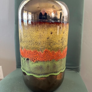 May include: A cylindrical ceramic vase with a glossy, metallic gold top and a dark brown base. The body features bands of yellow, orange, and green with a textured, speckled appearance. The vase is sitting on a green chair.