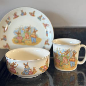 May include: A cream-colored ceramic dish set featuring a whimsical bunny design. The set includes a plate, a bowl, and a mug. The bunny illustrations are in pastel colors, creating a charming and festive aesthetic.