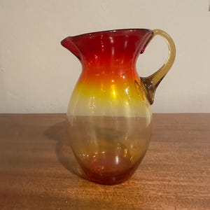 May include: A hand-blown glass pitcher with a gradient of colors. The top is a vibrant red, transitioning to yellow and then a clear base. It has a rounded body, a spout, and a curved handle. The pitcher is likely used for serving beverages.