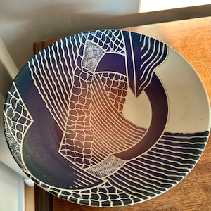 May include: A decorative ceramic plate with a unique abstract design. The plate features a gradient of colors, including deep blue, brown, and white, with intricate white line patterns. The design is reminiscent of a stylized landscape or abstract art.