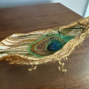May include: An elongated, boat-shaped decorative dish featuring a peacock feather pattern in shades of green, blue, and brown. The exterior and rim are finished in a textured gold leaf, and the piece is supported by three small, ornate gold legs.