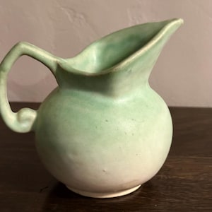 May include: A small ceramic pitcher with a rounded body and a curved handle. The exterior features a matte finish with a gradient of soft mint green at the top that fades into a creamy off-white color toward the base, including a pouring spout.