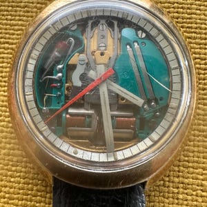 May include: A round gold-tone wristwatch with a black leather strap. The watch face features a transparent design revealing internal green and metallic components, with a red second hand and white hour and minute hands on a white dial ring.
