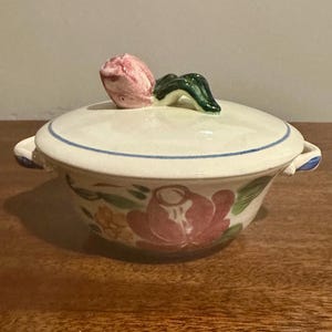 May include: A cream-colored ceramic lidded bowl featuring a hand-painted floral motif with pink and yellow flowers and green leaves. The lid is topped with a sculpted pink tulip handle and features a thin blue circular rim accent.