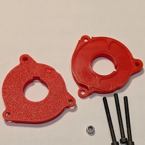 以下が含まれることがあります： 取り付け穴のある2つの赤い円形3Dプリントコンポーネントと、3本の黒いネジとナット。コンポーネントはテクスチャ加工された表面と中央の開口部を備えています。ハードウェアは白い表面に配置されています。