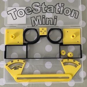 May include: A ToeStation Mini kit in a clear plastic storage case. The set includes yellow and black plastic components, including two yellow angle gauges with printed scales from 0 to 40, two yellow square plates, two black rings, and two small yellow knobs.