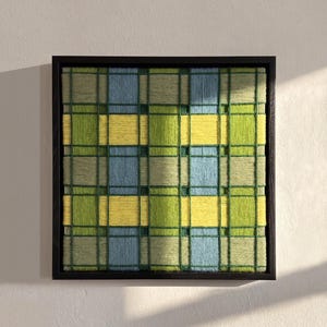 May include: A square wall art piece featuring a woven yarn design in a plaid pattern. The textile art displays vertical and horizontal bands of light blue, bright yellow, and various shades of green, all encased within a thin black wooden frame.