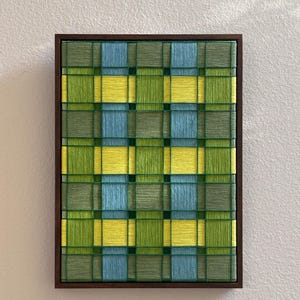 May include: A rectangular wall art piece featuring a woven grid pattern of yarn in shades of sage green, bright yellow, and light blue. The threads are layered over a dark frame, creating a textured, geometric design with intersecting lines.