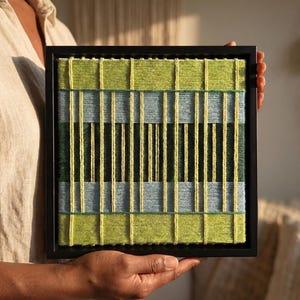 May include: A square wall art piece featuring a woven textile design in a black frame. The artwork consists of horizontal bands of lime green, light blue, and dark forest green yarn, overlaid with vertical strands of bright yellow yarn.
