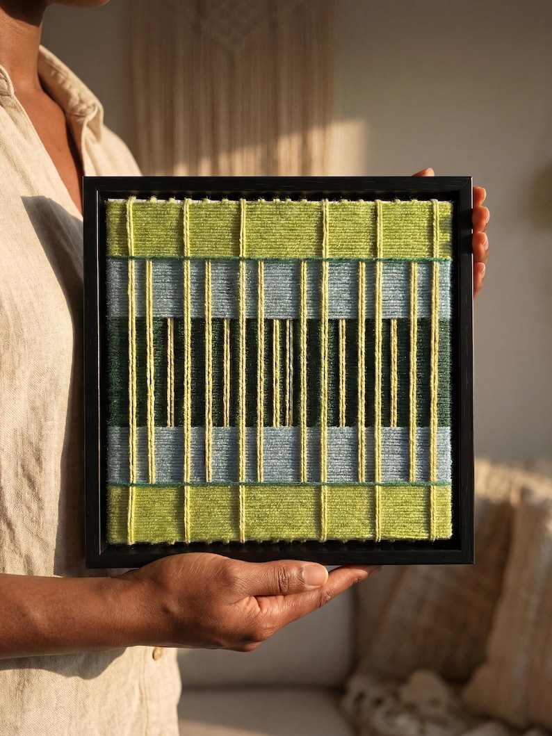 May include: A square wall art piece featuring a woven textile design in a black frame. The artwork consists of horizontal bands of lime green, light blue, and dark forest green yarn, overlaid with vertical strands of bright yellow yarn.