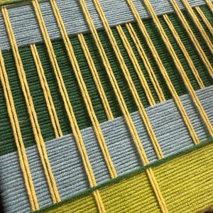 May include: A woven wall hanging featuring a geometric pattern of yarn in shades of lime green, forest green, and light blue. The design consists of horizontal bands of color intersected by vertical strands of bright yellow yarn throughout.