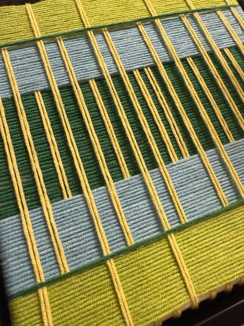May include: A woven wall hanging featuring a geometric pattern of yarn in shades of lime green, forest green, and light blue. The design consists of horizontal bands of color intersected by vertical strands of bright yellow yarn throughout.