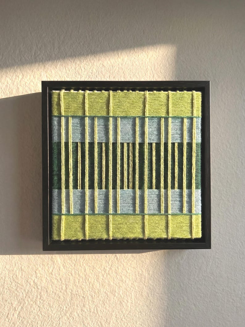 May include: A square wall art piece featuring a dark brown frame containing a woven textile design. The artwork consists of horizontal bands in lime green, light blue, and dark forest green, overlaid with vertical yellow yarn strands.
