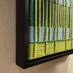 May include: A framed textile wall art piece featuring a woven grid pattern. The design consists of vertical and horizontal yarn strands in shades of lime green, forest green, and light blue, all encased within a simple black wooden frame.