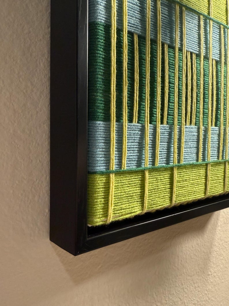 May include: A framed textile wall art piece featuring a woven grid pattern. The design consists of vertical and horizontal yarn strands in shades of lime green, forest green, and light blue, all encased within a simple black wooden frame.