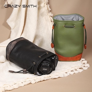 May include: A black leather drawstring camera bag containing a Sony camera lens and an upright green leather drawstring bag with a brown base. Both bags feature gold-tone hardware and cord closures. The brand name CRAZY SMITH is visible.
