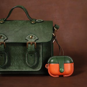 May include: A dark green satchel bag with a top handle, gold-toned buckles, and white contrast stitching, accompanied by a small, two-toned green and orange protective case for wireless earbuds featuring a snap button closure and white stitching.