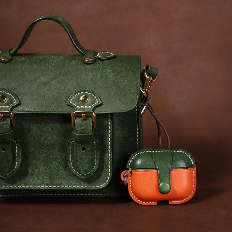 May include: A dark green satchel bag with a top handle, gold-toned buckles, and white contrast stitching, accompanied by a small, two-toned green and orange protective case for wireless earbuds featuring a snap button closure and white stitching.