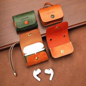 May include: Four leather cases for wireless earbuds, including two closed cases in green and tan, and two open cases in tan and green with white interiors. Each case features a snap button closure and the embossed text CRAZY CLOTH. Two white earbuds are placed below.