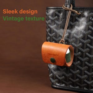 May include: A tan leather case with a dark green button closure and the text CRAZY SMITH embossed on the front, hanging from a black patterned tote bag by a thin brown cord. The text Sleek design and Vintage texture appear in the image.