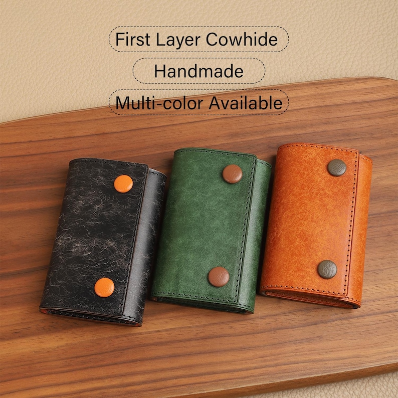 May include: Three leather key wallets in black, green, and orange, each with a button closure. The wallets are displayed on a wooden surface. Text on the image reads: "First Layer Cowhide", "Handmade", and "Multi-color Available".