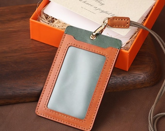 May include: A brown leather ID badge holder with a green top section and a clear window. The holder is attached to a lanyard. A gift box with a card that reads "May You have a joyful life" is in the background.