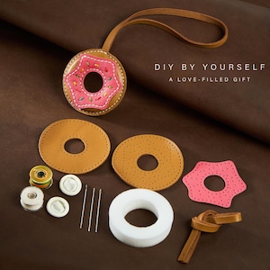 May include: DIY donut-shaped leather keychain kit. The kit includes pre-cut leather pieces in brown and pink, thread, needles, and a white foam insert. The finished keychain features a pink donut with sprinkles and a brown leather strap.