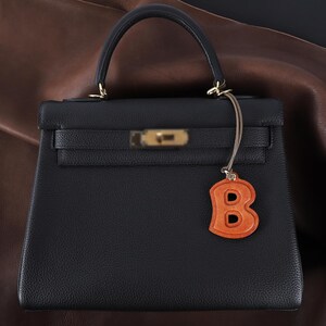 May include: A structured black handbag with a single top handle, a gold-toned metal clasp closure, and a textured leather finish. An orange leather charm in the shape of the letter B hangs from a thin cord attached to the handle hardware.