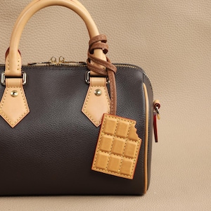 May include: A brown leather handbag with a tan leather strap and a chocolate bar-shaped charm. The bag has a zipper closure and gold hardware.