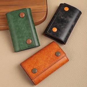 May include: Three leather key holders in green, black, and orange. Each key holder is rectangular with a snap closure and a contrasting button. The key holders are arranged on a light brown surface.