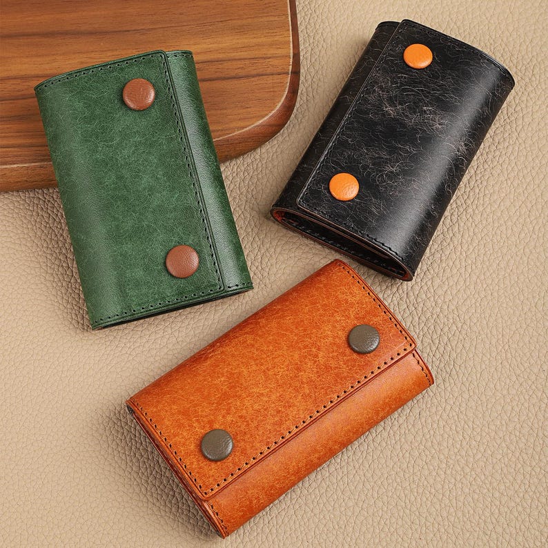 May include: Three leather key holders in green, black, and orange. Each key holder is rectangular with a snap closure and a contrasting button. The key holders are arranged on a light brown surface.