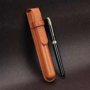 May include: A black fountain pen with gold accents rests beside a brown leather pen case. The pen case is rectangular with a flap and a strap closure. The pen has a sleek design, and the case has a textured surface.