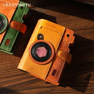 May include: A yellow and orange leather wallet designed to resemble a vintage camera, featuring a circular black lens detail with a hexagonal center, a snap closure, and the text CRAZYSMITH embossed on the top front surface.